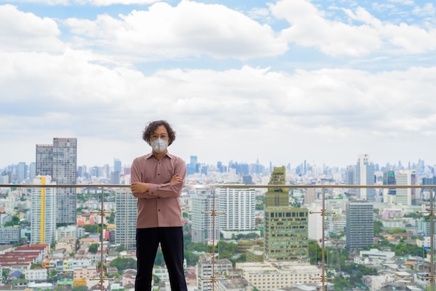 Japanse zakenman met krullend haar draagt masker ter bescherming tegen uitbraak van coronavirus tegen uitzicht op de stad