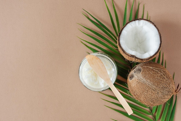 Jar of coconut butter and fresh coconuts with palm leaves on brown background top view