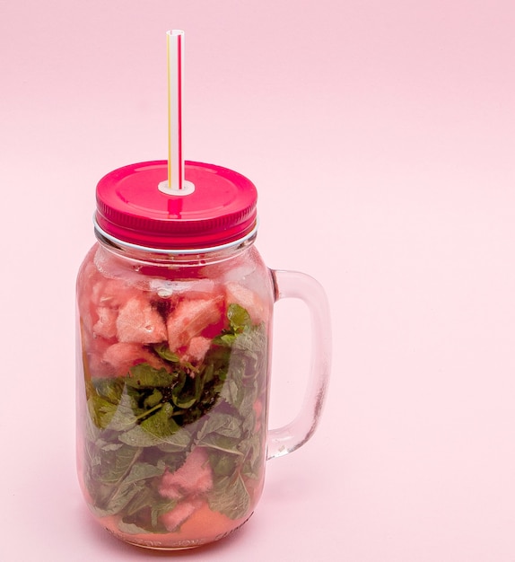 Jar of cold fresh lemonade with piece of watermelon and drinking straws on pink wall.