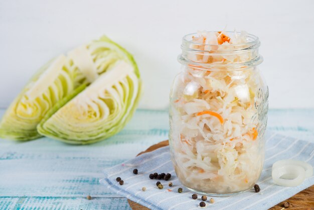 A jar of fermented cabbage stands on a wooden table to the right with fresh vegetables around. Horizontal orientation with copy space.