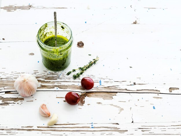 Jar of fresh home made pesto with garlic cherrytomatoes and thyme over rustic white wooden backdrop Top view