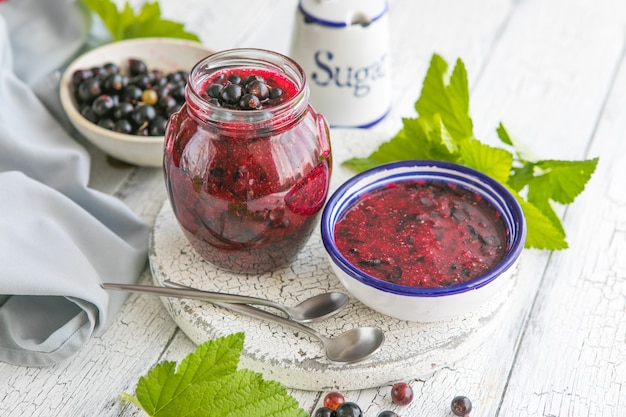 Jar of homemade fresh currant jam with shugar fresh berries\
black currant on white wooden background