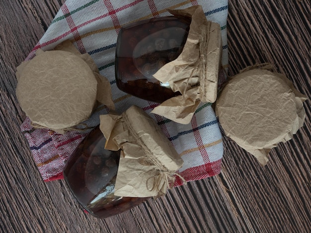 A jar of homemade strawberry jam, covered with parchment, lies on a wooden background