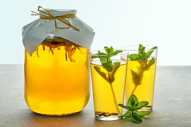 Jar of kombucha and two glass glasses of kombucha are garnished with mint and lime lemon on a light gray concrete table