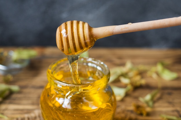 A jar of liquid honey from Linden flowers and a stick with honey on a dark surface.