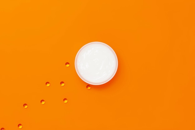 오렌지 배경에 보습 화이트 크림 한 병 옆에 비타민 E가 들어있는 캡슐이 있습니다.