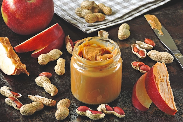 Jar of peanut butter, peanuts in a peel, apple slices and a knife