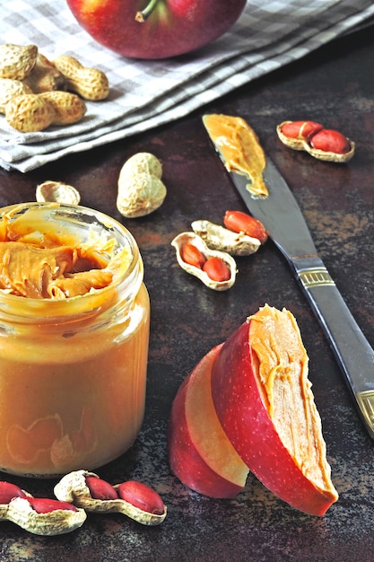 Jar of peanut butter, peanuts in a peel, apple slices and a knife