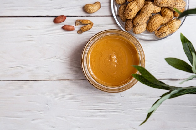 A jar of peanut butter with a bunch of peanuts on a white wooden table with an olive branch