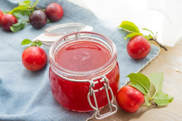 A jar of plum jam sits on a blue cloth next to a bunch of cherries.