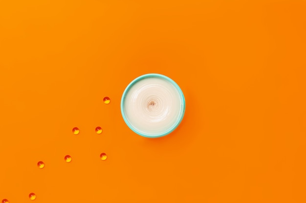 A jar of white clay on an orange background next to which are capsules with vitamin E.