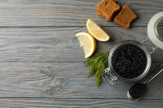 Jar with black caviar, bread, dill and lemon on wooden background, space for text