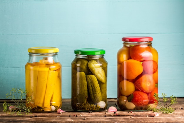 Jars with pickles of courgettes, cucumbers, tomatoes on a blue, wooden background.