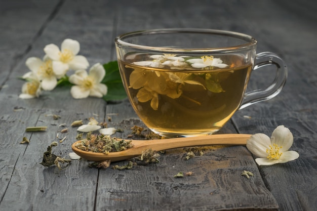 Foto fiori di gelsomino e tè ai fiori in una ciotola di vetro su un tavolo di legno. una tazza di tè ai fiori e un cucchiaio di legno con erbe secche.