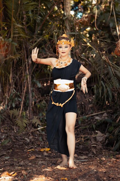 Javanese woman dancing pose in a black tank top and black skirt with golden crown and golden accessories on her body inside the jungle