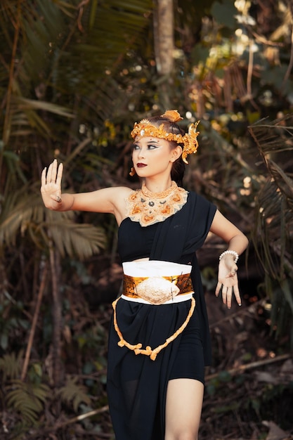 Javanese woman dancing pose in a black tank top and black skirt with golden crown and golden accessories on her body inside the jungle