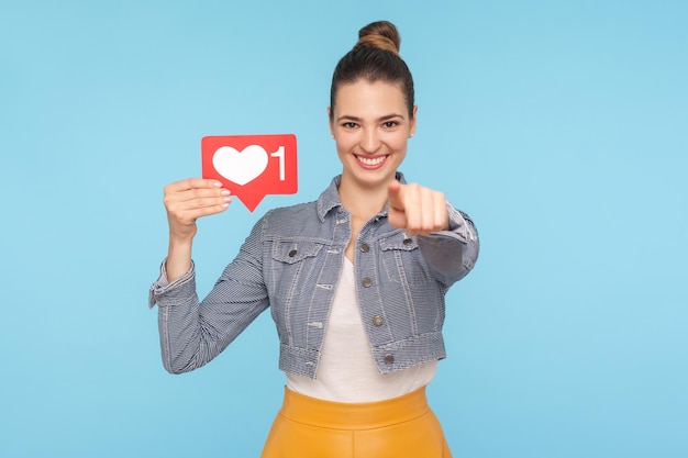 Je duwt op de Like-knop van het netwerk Portret van een positieve, modieus geklede vrouw met een haarknot die het hartpictogram van de sociale media vasthoudt en naar de camera wijst die aanbeveelt om de studio-opname van de liefdesinhoud te volgen