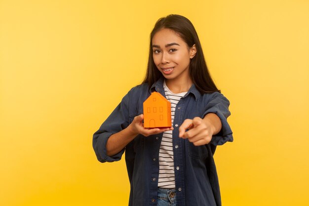 Je eigen toekomstige huis. Portret van een vrolijk meisje in een spijkerblouse die een papieren huis vasthoudt en naar de camera wijst, aanmoedigt om onroerend goed te kopen, een gunstige hypotheek te nemen. indoor studio-opname, geïsoleerd