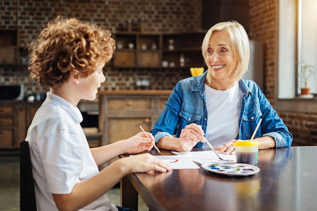 Je maakt een grapje. Nadenkende oudere vrouw breed grijnzend terwijl ze schilderde met haar kleinkind en luisterde naar zijn verhalen aan een familietafel.