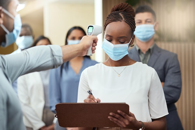 Je temperatuur is Bijgesneden opname van een aantrekkelijke jonge zakenvrouw die een masker draagt en door het covid-scherm gaat terwijl ze aan het hoofd van een rij in haar kantoor staat