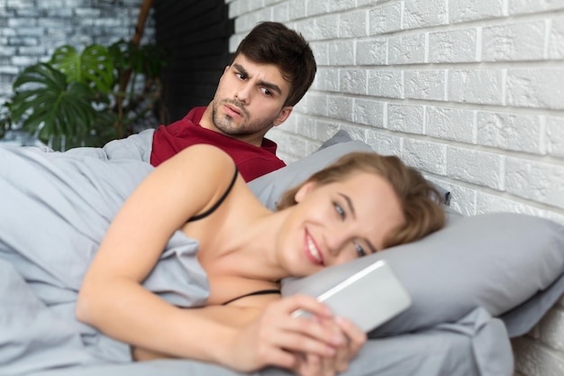 Jealous boyfriend. Shocked man looking at girlfriends mobile phone while she reading message, lying in bed