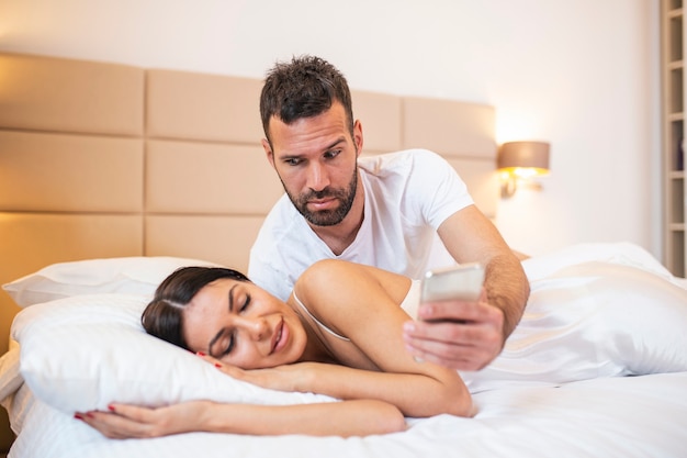 Jealous husband spying the phone of his partner while she is sleeping in a bed at home.