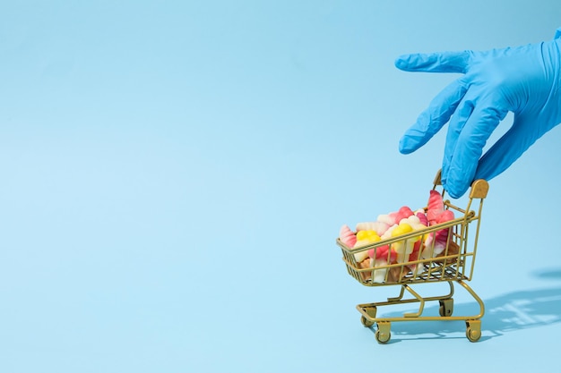 Jelly candies in shopping trolley and hand in medical glove on blue background space for text