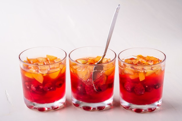 Jelly dessert with fruits apricot raspberry in three drinking glasses with single spoon yellow red summer white surface isolated in row