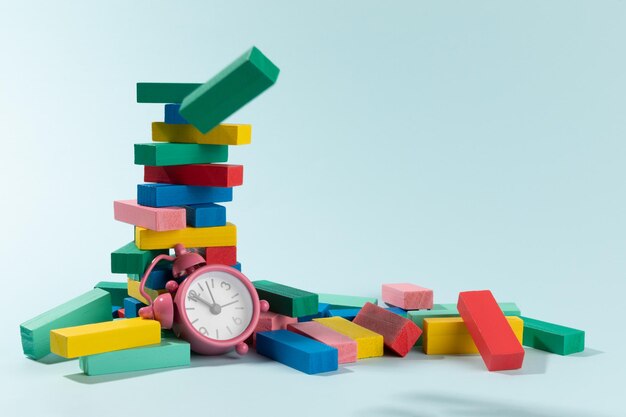 Jenga tower made of color wooden blocks and a pink clock on light blue background