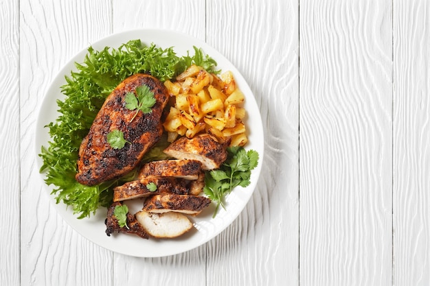 Jerk chicken breasts with grilled ananas cubes and fresh green leaves lettuce on a white plate on a wooden textured table horizontal view from above flat lay free space