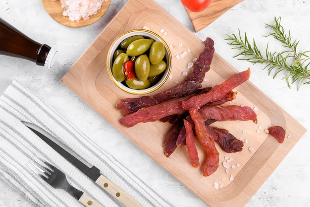 Jerky meat strips with spices, green olives, rosemary on a grey background.