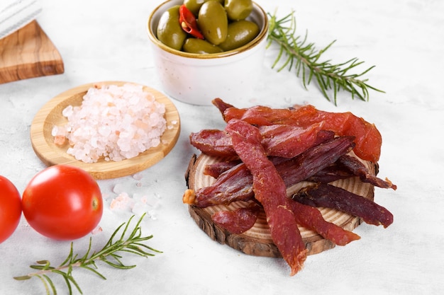 Jerky meat strips with spices, green olives, rosemary on wooden plate on a grey stone background. Tipical Italian food coppiette is a spiced pork meat strips. Snacks for beer, close up