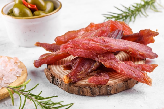 Jerky meat strips with spices, green olives, rosemary on wooden
plate on a grey stone background. tipical italian food coppiette is
a spiced pork meat strips. snacks for beer, close up