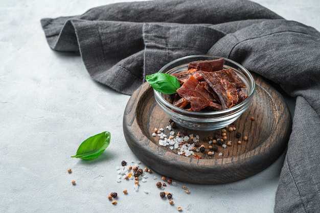 Jerky meat on a wooden plate with spices, salt and basil on a gray wall. Side view with copy space.