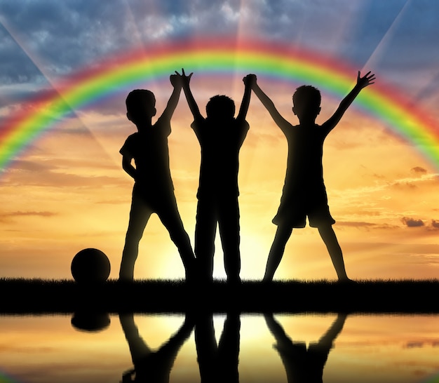 Jeugd concept. Drie gelukkige kinderen hand in hand op een regenboog achtergrond en de zonsondergang boven het water met hun reflectie