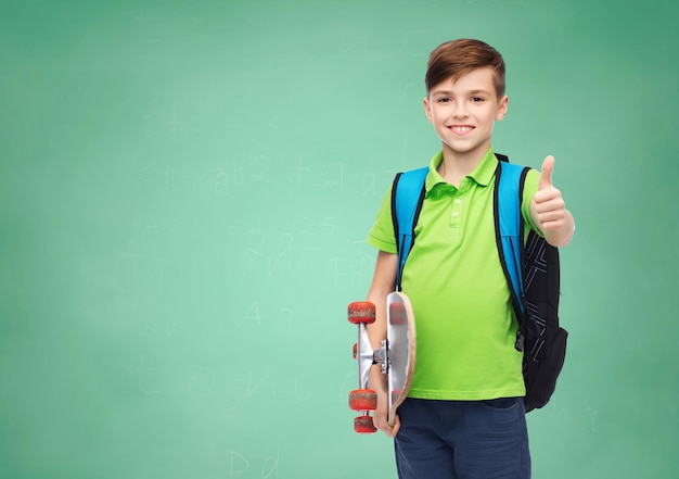 jeugd, gebaar, onderwijs en mensen concept - gelukkig lachende student jongen met rugzak en skateboard duimen opdagen over groene school krijtbord achtergrond