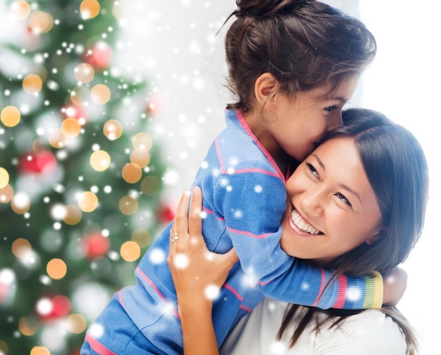 jeugd, geluk, kerst, familie en mensen concept - glimlachend klein meisje en moeder knuffelen binnenshuis over woonkamer met boom