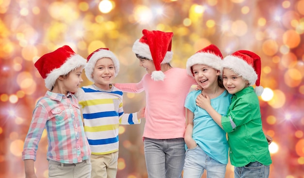 jeugd, kerstmis, vakantie, vriendschap en mensenconcept - groep gelukkige lachende kleine kinderen in kerstmutsen knuffelen over de achtergrond van vakantieverlichting