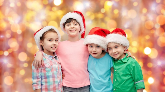 jeugd, kerstmis, vakantie, vriendschap en mensenconcept - groep gelukkige lachende kleine kinderen in kerstmutsen knuffelen over de achtergrond van vakantieverlichting
