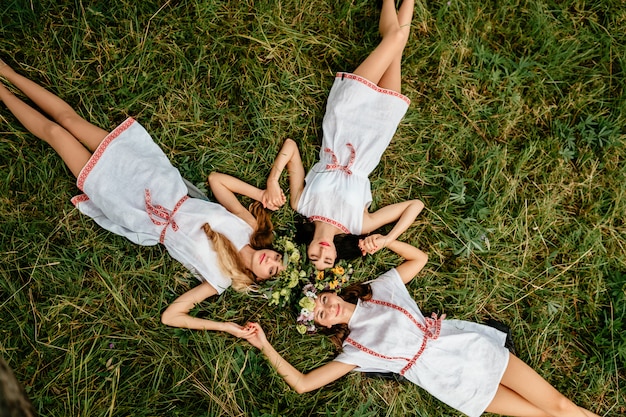 Jeugd levensstijl. Drie verbazingwekkende Slavische etnische meisjes van de verschijnings etnische volksstijl met kroon die van bloemen op gras onder boom bij aard in de zomer liggen.