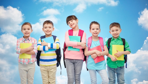 Foto jeugd, voorschoolse educatie, leren en mensen concept - groep gelukkige lachende kleine kinderen met schooltassen en notitieboekjes over blauwe lucht en wolken achtergrond