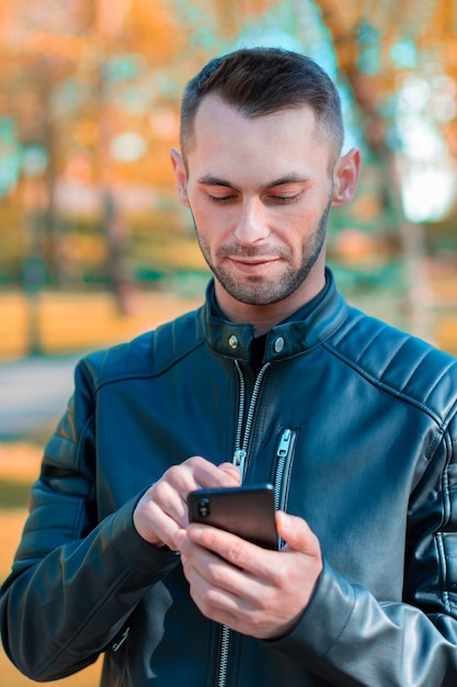 Jeugdige man met zwarte Smartphone in het prachtige herfstpark. Knappe jongeman met mobiele telefoon op zonnige dag - medium close-up shot