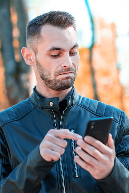 Foto jeugdige man was verrast met behulp van zwarte smartphone in het prachtige herfstpark. knappe jongeman met mobiele telefoon op zonnige dag - medium close-up portret