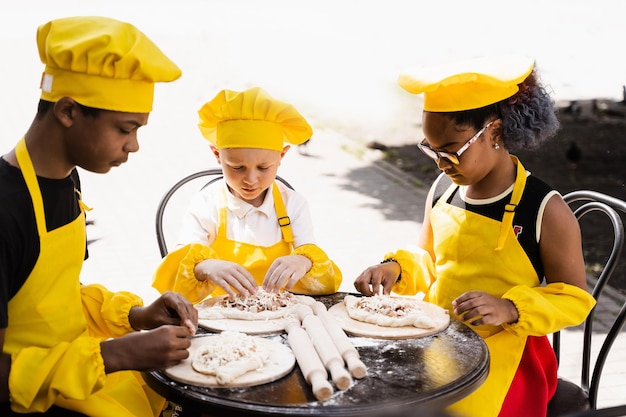 Jeugdkokactiviteit van multinationale kinderen van zwarte Afrikaanse en blanke kinderen in gele koksmuts en schortuniform Gelukkig gezelschap van multi-etnische kinderen die deeg koken