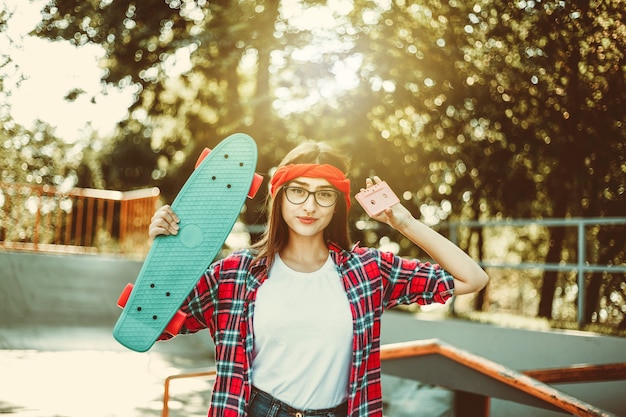 Jeugdlook in retrostijl. jaren 80. Jonge, aantrekkelijke hipstervrouw in stijlvolle kleding houdt skateboard en audiocassette vast terwijl ze op een zonnige dag voor de camera in het skatepark poseert