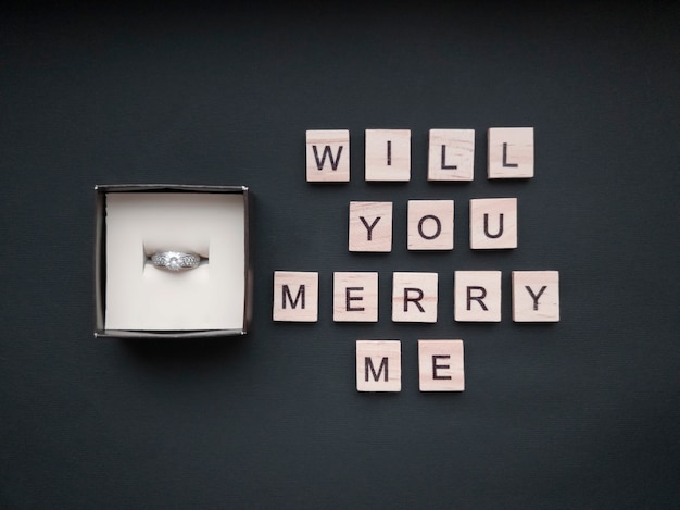 In the jewelry box there is a ring with a stone and an inscription from wooden squares will you marry me on a beautiful black background. Romantic concept. Flat style.