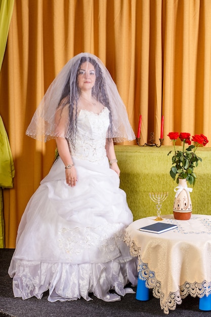 Una sposa ebrea in abito bianco, la testa coperta da un velo, sta in una stanza a un tavolo con fiori, aspettando lo sposo per la cerimonia della chuppah