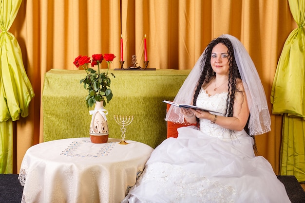 A Jewish bride in a white wedding dress with a veil sits at a table with flowers and reads blessings from a prayer book before the chuppah ceremony