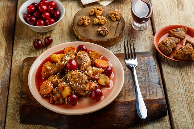 Jewish dish stewed potatoes with chicken in cherry sauce decorated with cherries on a stand next to a fork and ingredients.