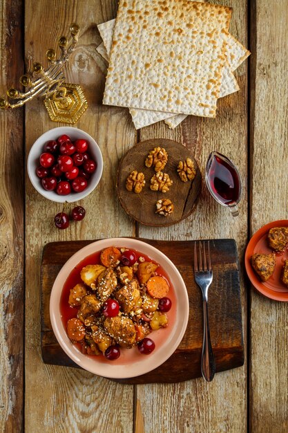 Jewish dish stewed potatoes with chicken in cherry sauce decorated with cherries on the table in a plate next to matzo and menorah. Vertical photo